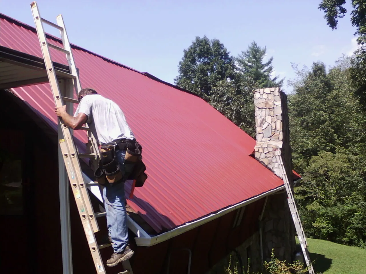 Roofer on a ladder beside a red metal roof and stone chimney — Soffit & Fascia Repair by Roofing Brothers in College Springs